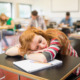 Syudent sleep depravation: Reen redhead girl sleeps with hed on arms sitting at classroom vesk surrounded ny other students at desks