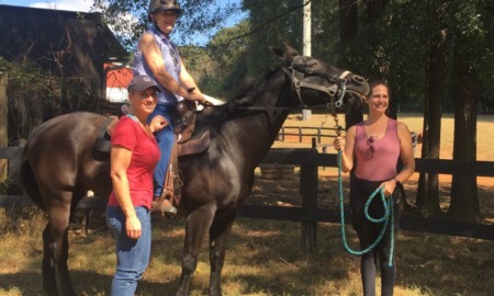 Disability rights: A smiling woman sits atop a dark brown horse. Two other smiling women stand on the ground on either side of the woman on the horse.