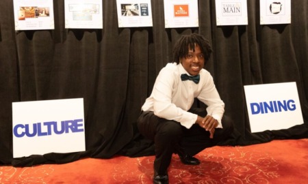 Atlanta student homelessness: Young Black man with black dreads in formal wear with black bow tie crouches on orange carpet in front of black curtain with several hanging paper signs.