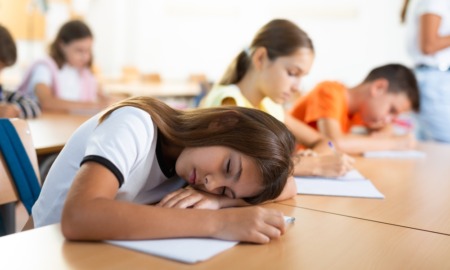 Student Overscheduling: Tired, dark-haired, preteen schoolgirl sleeping at desk in classroom during lesson with her head resting on her hands