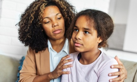 social and emotional learning, SEL: Black teen girl with brown hair in white top sits next to Black woman with brown hair in tan jacket and white top on light couch. Woman holds girl around shoulders.