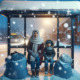 Homelessness in Massachusetts: Woman and young boy in heavy winter clothing sit at covered city bus stop at night in heavy snow with two large cloth bags at their feet