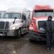 Prison No Education: Black man in dark winter jacket, hat and pants stands leaning against front of bright red semi-truck parked next to a white semi-truck on asphalt parking lot under gray, rainy sky