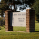 Q&A Teaching after Columbine: Columbine High School sign - cement center with red brick pillars - in 2012, on grass slope with evergreen trees in background.