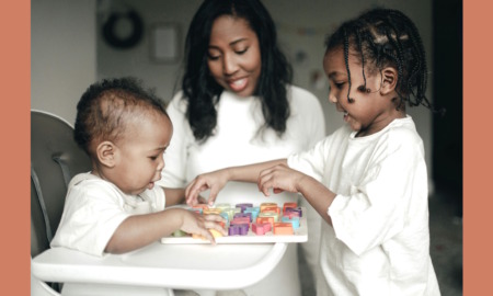 Guaranteed Income: Black mother, infant in a white high chair and toddler — all dressed in white — play with colorful 3D letter on a puzzle board on the high chair tray