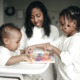 Guaranteed Income: Black mother, infant in a white high chair and toddler — all dressed in white — play with colorful 3D letter on a puzzle board on the high chair tray