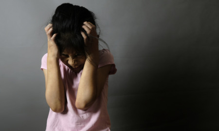 Suspension disabled students: young girl with dark hair in pink r-shirts holds head in hands with angry face against shadowed, dark background