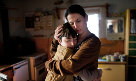 Child support payments: Portrait of sad poor mature mother with dark hair pulled back in brown sweater, standing hugging preteen daughter indoors at home in older, dark kitchen.