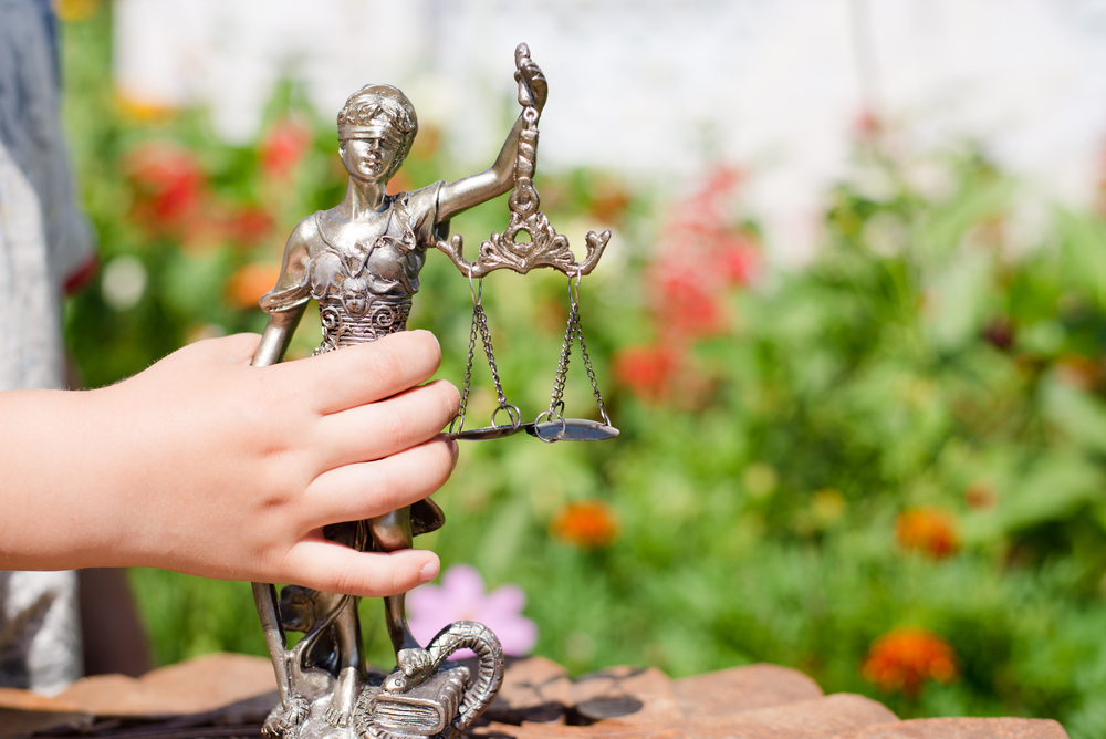 Legal counseling for behavior issues: Young child's hand holding sculpture of themis, femida or justice goddess on green leaves natural bokeh copy space background