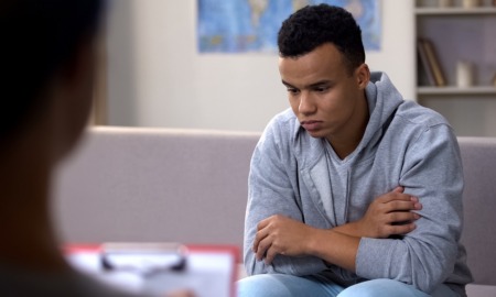 Community-youth-violence-prevention: Black teen sullenly sits on couch across from counselor