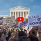 Abortion bans: Crowd of adults at rally with handmaid signs held up gathered in front of multi-story, white building with 8 pillars in front