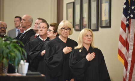 Oklahoma-strikes-down-religious-charter-school: Several people in black judge's robes stand at attention on interior steps with hand over heart facing American flag