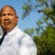 Uvalde indictment: Bald white man in white business shirt and dark tie stands staring seriously into camera under blue sky with green trees in the background.