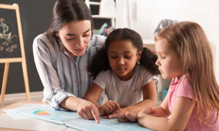 K-3-literacy-scores: Two young girls sit at desk with teacher reading a book.