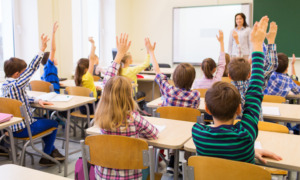 Segregation academies: Back view of group of all white school kids sitting at classroom desks and raising hands with white female teacher standing facing students