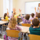 Segregation academies: Back view of group of all white school kids sitting at classroom desks and raising hands with white female teacher standing facing students