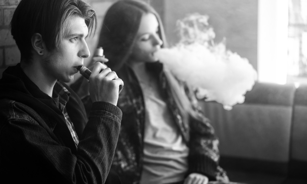 youth tobacco use, vaping: Black and white photo of two older teens sitting against brick wall smoking electronic cigarettes