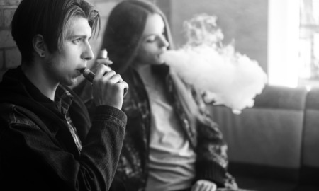 youth tobacco use, vaping: Black and white photo of two older teens sitting against brick wall smoking electronic cigarettes