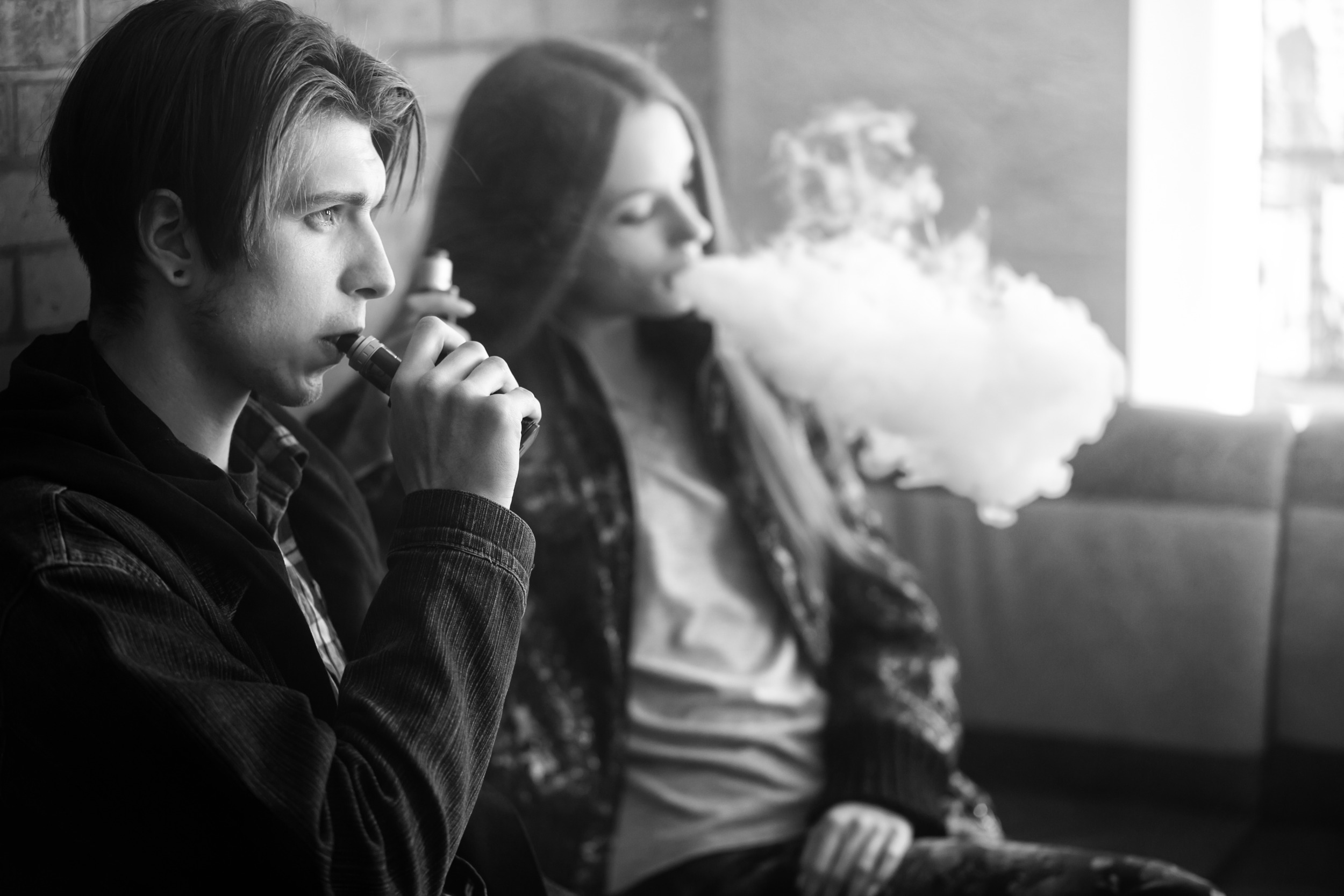 youth tobacco use, vaping: Black and white photo of two older teens sitting against brick wall smoking electronic cigarettes