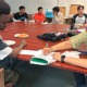 Older Immigrant students high school enrollment: Group of Black, male, older teens sit stound wood conference table with older brown-haired white woman helping one in foreground to fill out paperwork