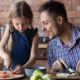 Math talk by parents to children: Adult man with short brown hair in blue plaid shirt talks to young girl with long blonde hair in navy dress as both stand at kitchen counter chopping vegetables