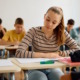 Reading literacy: Teen girl with long blonde hair in ponytail sits in classroom reading book on her desk with other students in background reading at their desks