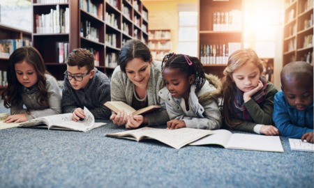 Book Ban Survey: Several young elementary students with one adult female teacher lie on library blue rug reading books in front of bookshelves filled with books