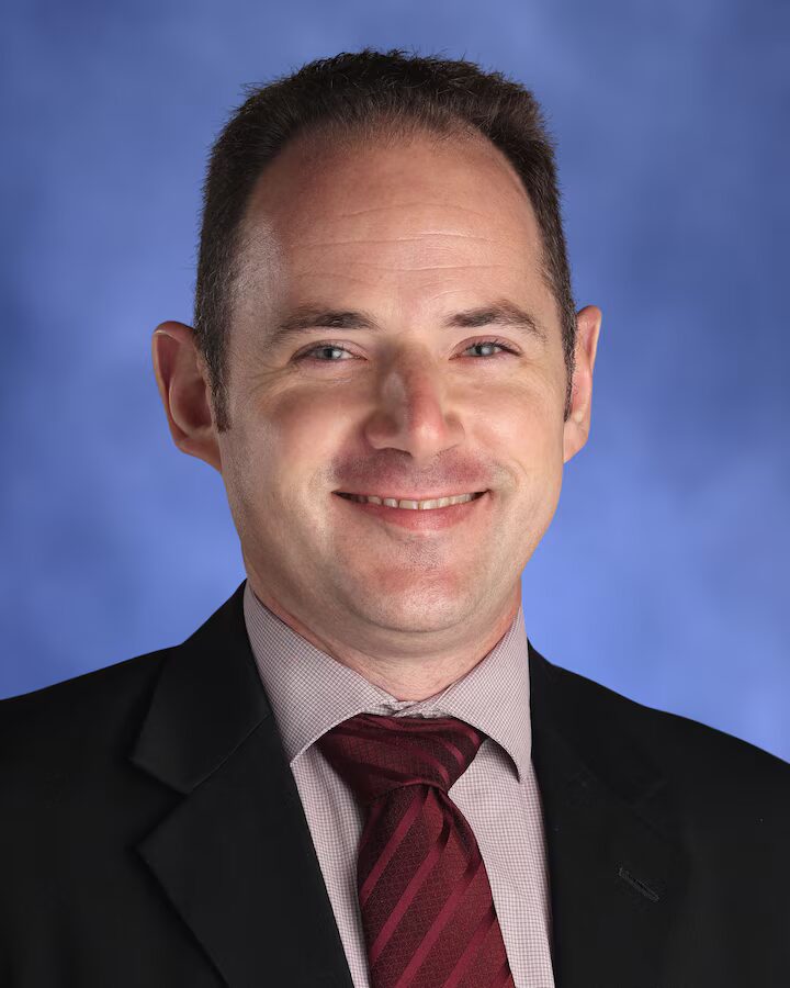 CIVICS TEACHER INTERVIEW: HEADSHOT MAN WITH SHORT DARK HAIR IN BLACK SUIT WITH TAN SHIRT AND BURGUNDY TIE