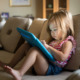 Preschooler excessive screen time study: Young girl in red top and blue shorts with long light brown hair sitting at home on tan couch and using a child's tablet touch screen computer