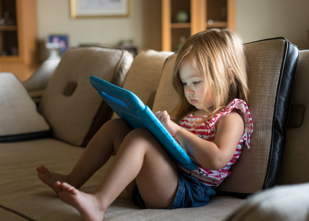 Preschooler excessive screen time study: Young girl in red top and blue shorts with long light brown hair sitting at home on tan couch and using a child's tablet touch screen computer