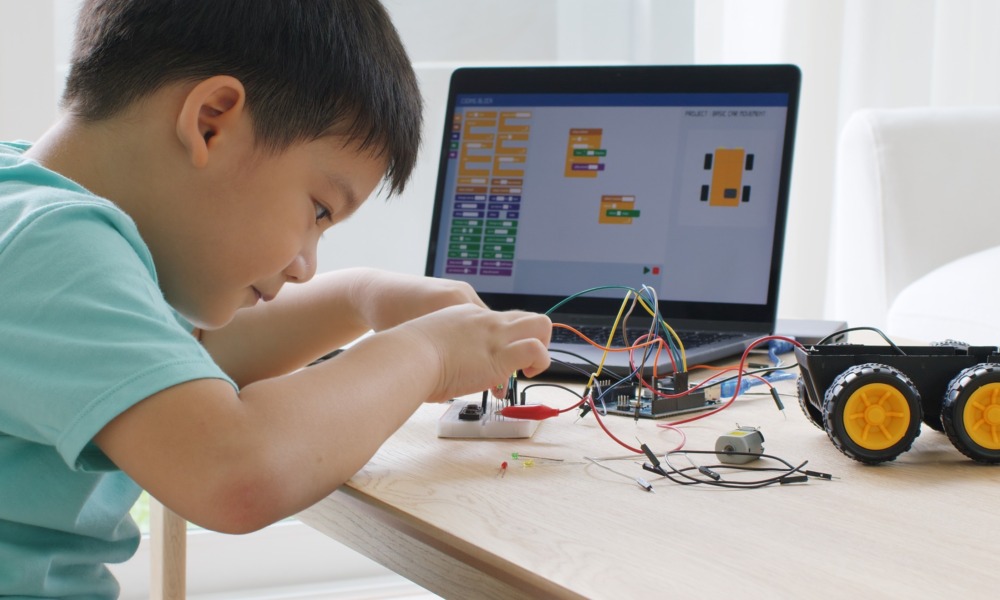 education opportunity gag: Young boy with dark hait sits at desk playing with wiring on robot next to a computer monitor