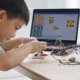 education opportunity gag: Young boy with dark hait sits at desk playing with wiring on robot next to a computer monitor