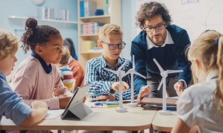 7 successful schools: Male teacher leans over table of 5 middle school students working on building models ofwind turbine power generators