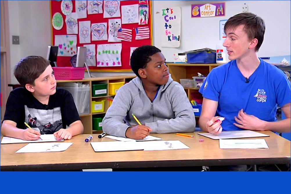 AMERICORPS TUTORS: YOUNG MAN IN BRIGHT BLUE AMERICOPRS POLO SHIRT SITS WITH TWO YOUNG BOYS IN CLASSROOM AT LONG DESK WRITING IN PAPER WORKBOOKS.