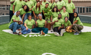 Restorative justice partnerships: Group of around 20 smiling people all wearing lime green t-shirts stand and sit in 3-4 rows on an artificial green turf field surrounded by tall city buidings