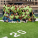 Restorative justice partnerships: Group of around 20 smiling people all wearing lime green t-shirts stand and sit in 3-4 rows on an artificial green turf field surrounded by tall city buidings