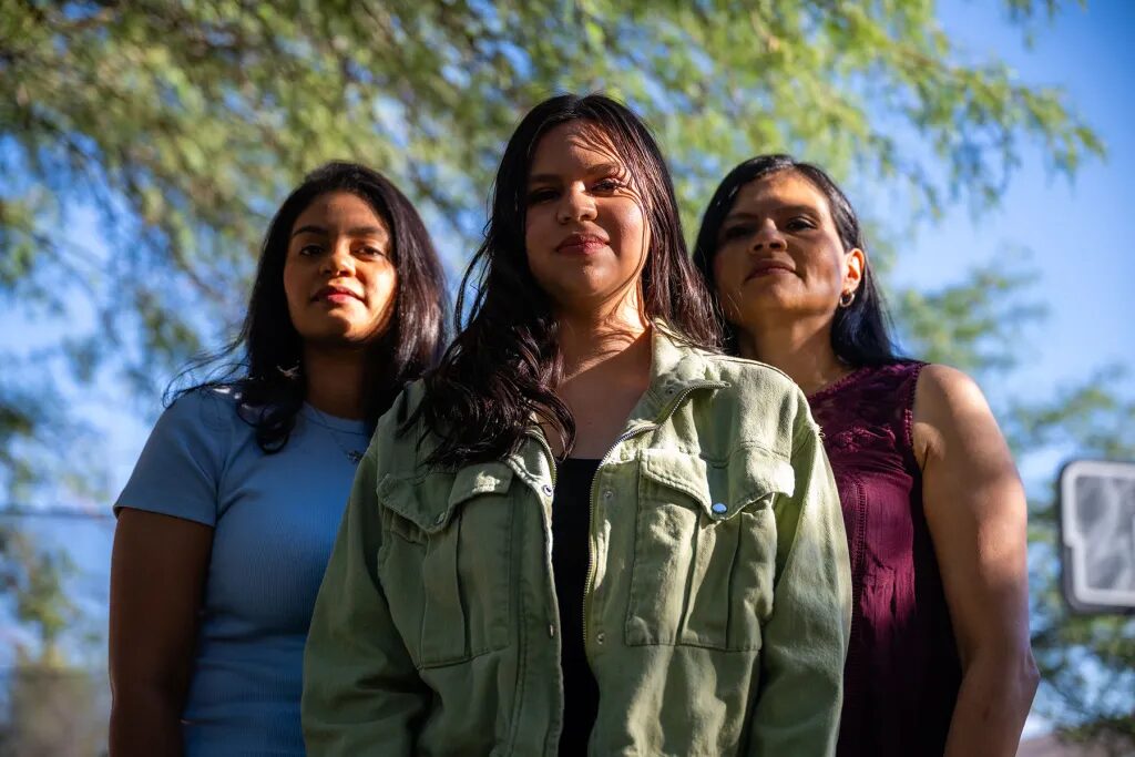 Latino gun reform Arizona: Thre Latino women with long, dark hair stand together facing camera with green tree foliage in the background