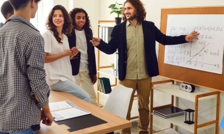 Mixed algebra class experiment: Group of young teens stand around display board watching male teacher in dark sport coat and tan pants explain an algebra formula on a white paper pad