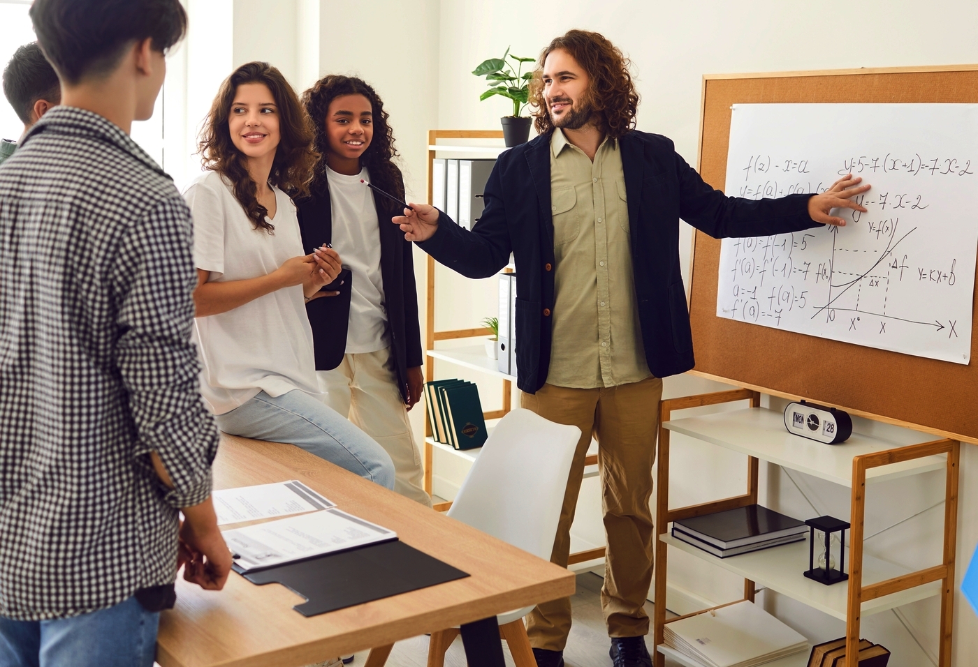 Mixed algebra class experiment: Group of young teens stand around display board watching male teacher in dark sport coat and tan pants explain an algebra formula on a white paper pad