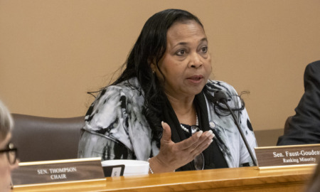 Foster care sleep-only placement: Black woman with long braids in black and white print jacket seated at table speaks into microphone gesturing with right hand