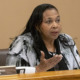 Foster care sleep-only placement: Black woman with long braids in black and white print jacket seated at table speaks into microphone gesturing with right hand