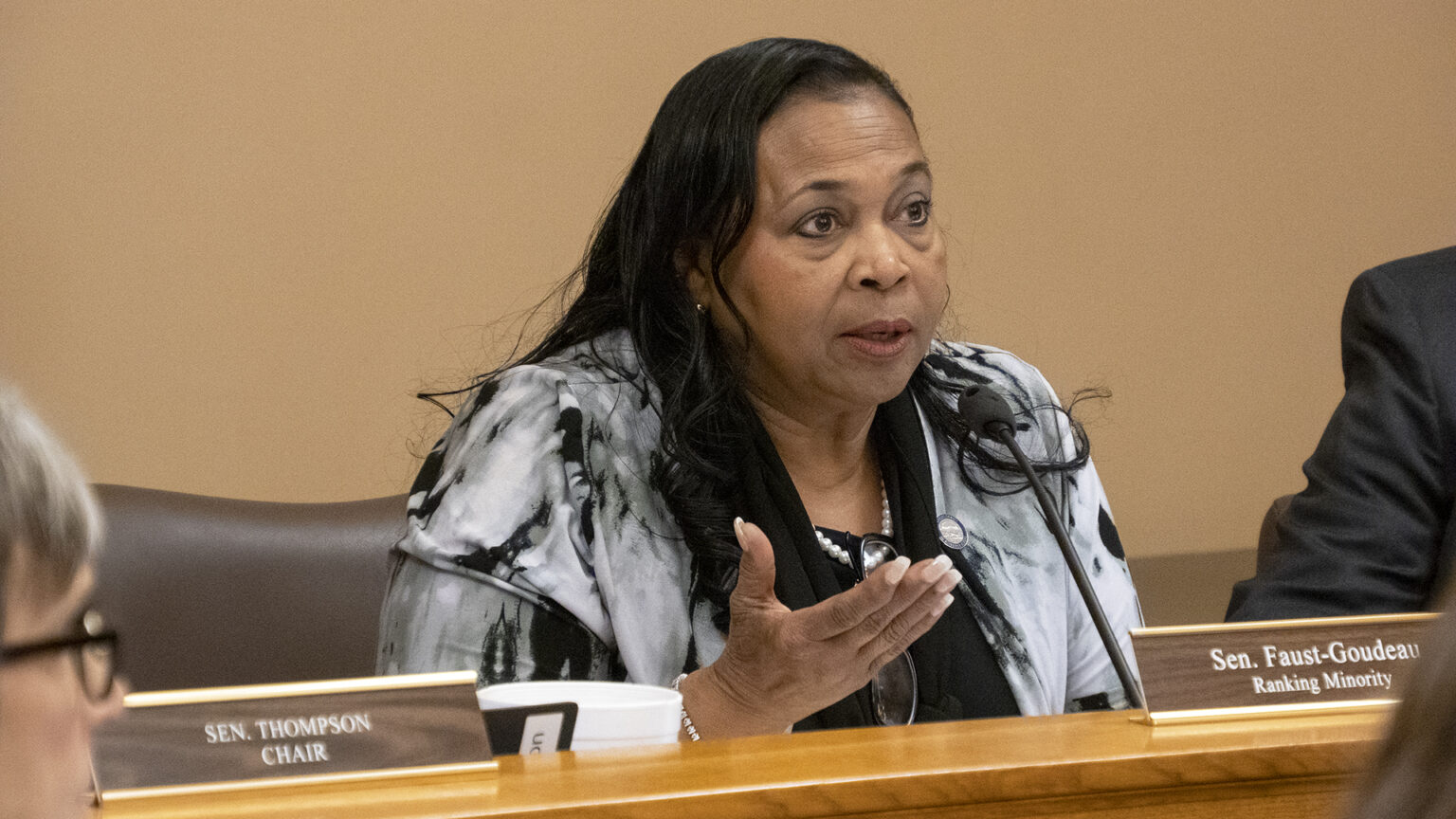 Foster care sleep-only placement: Black woman with long braids in black and white print jacket seated at table speaks into microphone gesturing with right hand