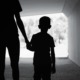 West Virginia foster care: Blac k and white photo of silhouette back of adult holding hand of young child, walking away from camera in dark tunnel toward light outdoor area