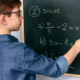 Algebra 8th grade options survey: Middle school age boy with short brown hair and glasses stands at chalkboard solving simple algebra equation