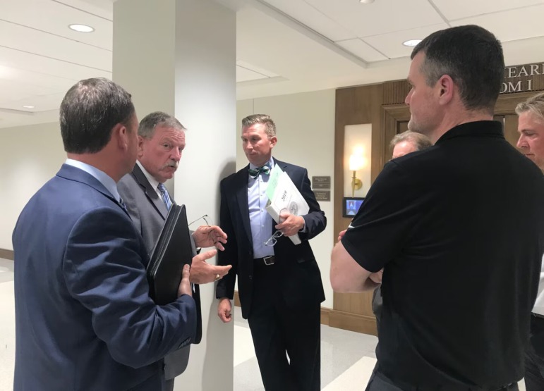 Arming teachers in Tennessee: Six middle-age men, all with gray or graying hair, in dark suits stand in white interior hallway talking