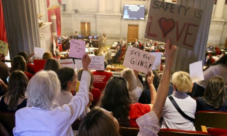 Arming teachers in Tennessee: Back view of several people holding up hand-printed signs seated in second story gallery overlooking government officials in session on first floor