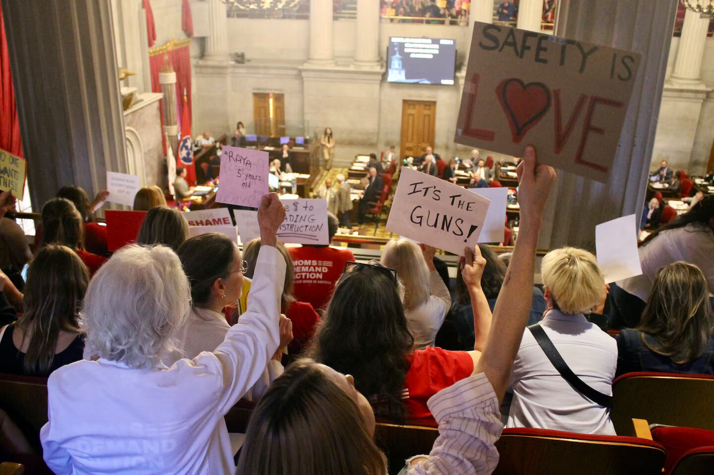 Arming teachers in Tennessee: Back view of several people holding up hand-printed signs seated in second story gallery overlooking government officials in session on first floor