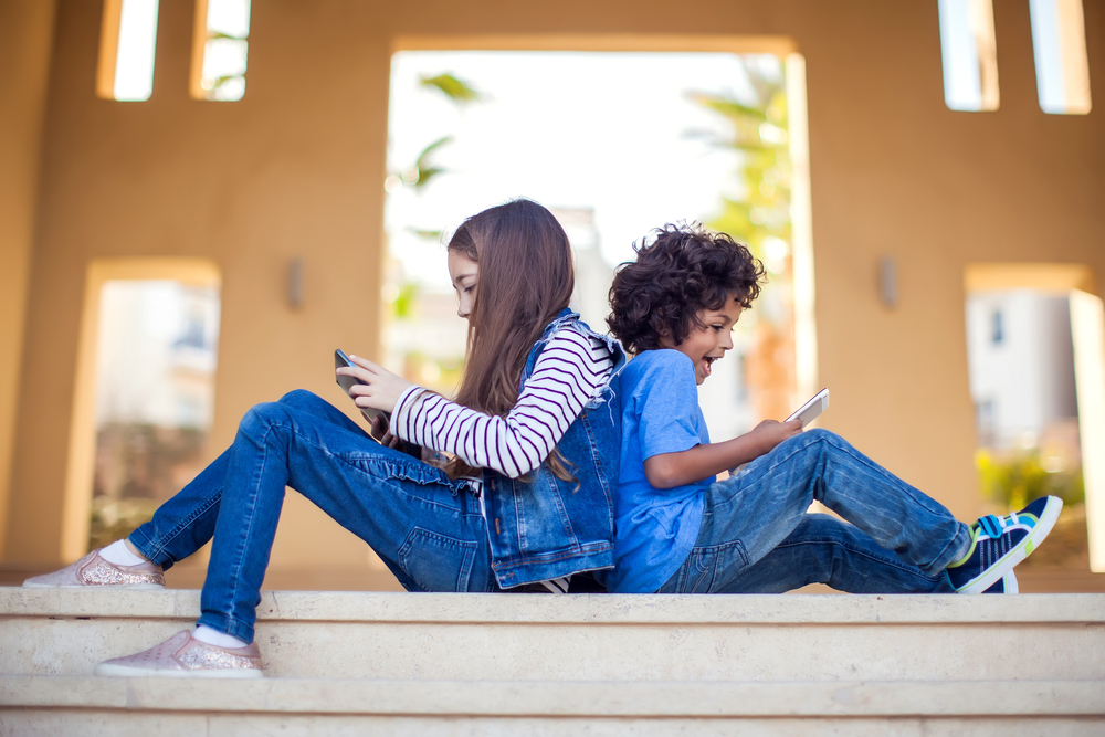 Q&A social media and kids: Two prettens sitting back to back on the floor in front of a window play games on tablets.