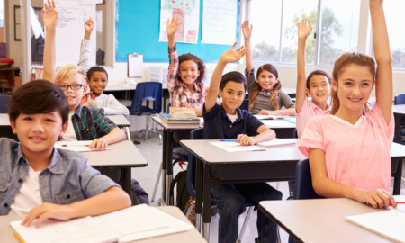 Pittman opinion education ecosystem: Elementary school kids in a classroom sitting at desks raising their hands