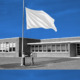 Civic curriculum debate: Two children face each other at very tall flagpole raising an all-white flag up the pole that is on a lawn in front of a one-story brick building with multiple large windows. Blue sky above building.